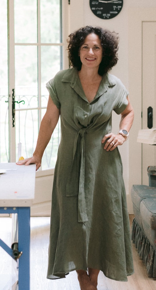 A photo of Dale standing by here main work table in the lantern cozies studio above the garage. Its a sunny afternoon and she's wearing a green linen dress