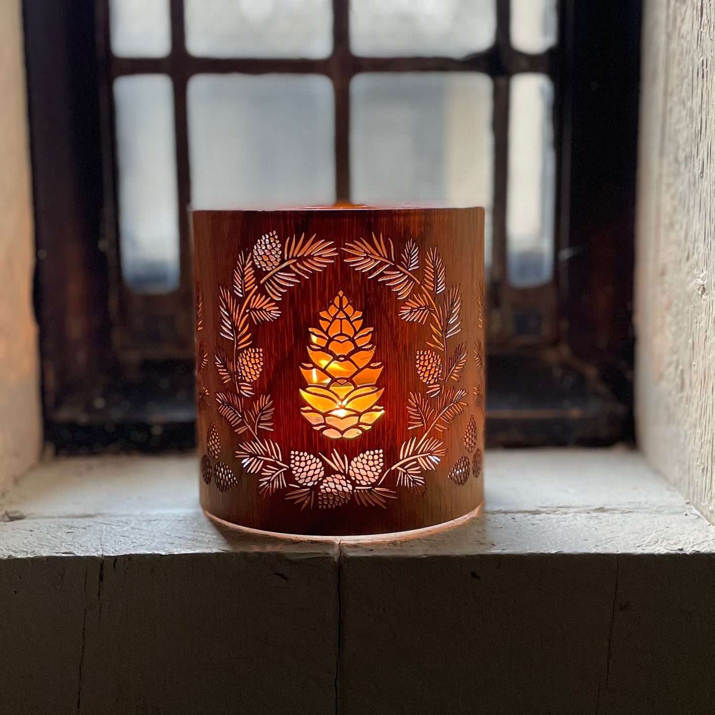 a glass lantern candle holder wrapped in white oak veneer with laser cut pinecone design lit with a candle