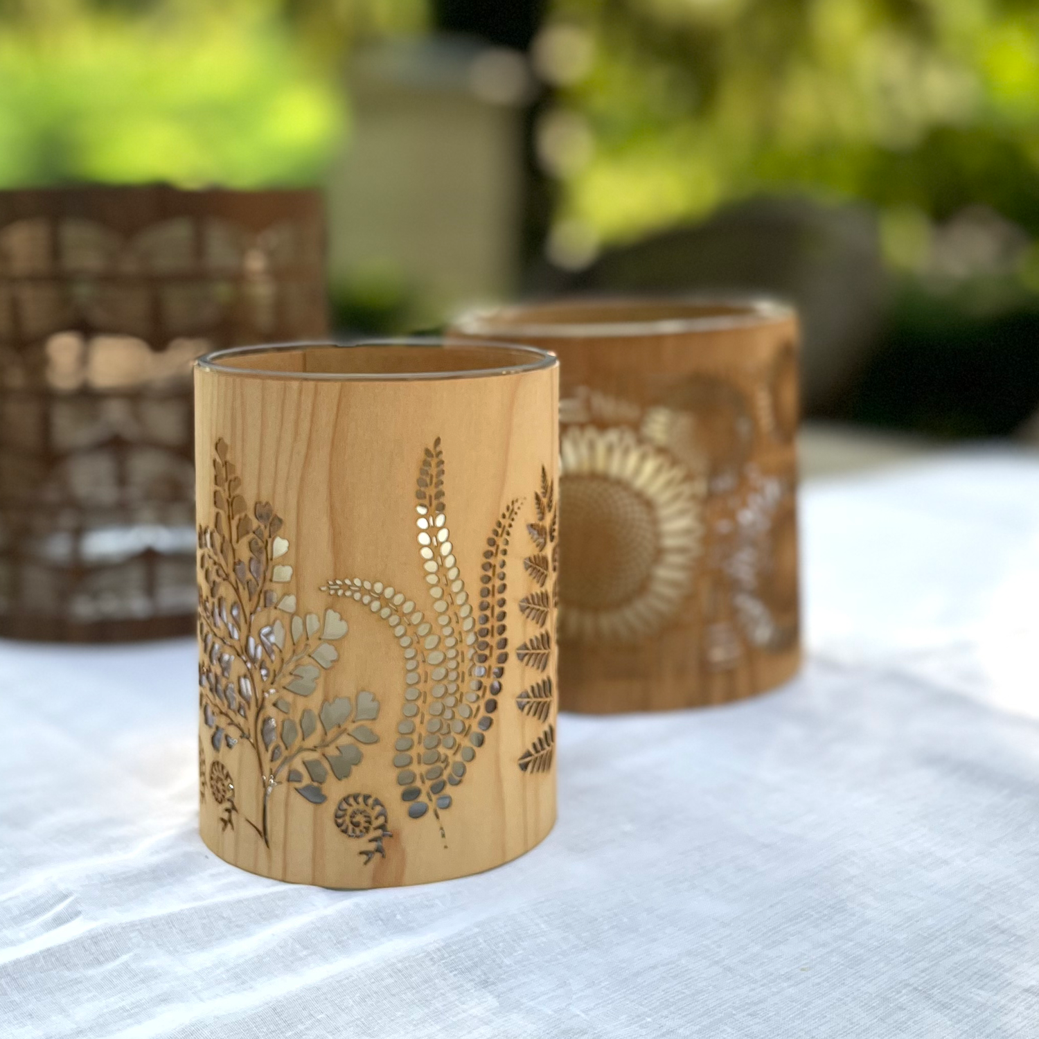 Two cylindrical wooden candle holders with laser-cut botanical and sunburst patterns, displayed on a white fabric surface with a blurred green outdoor background.