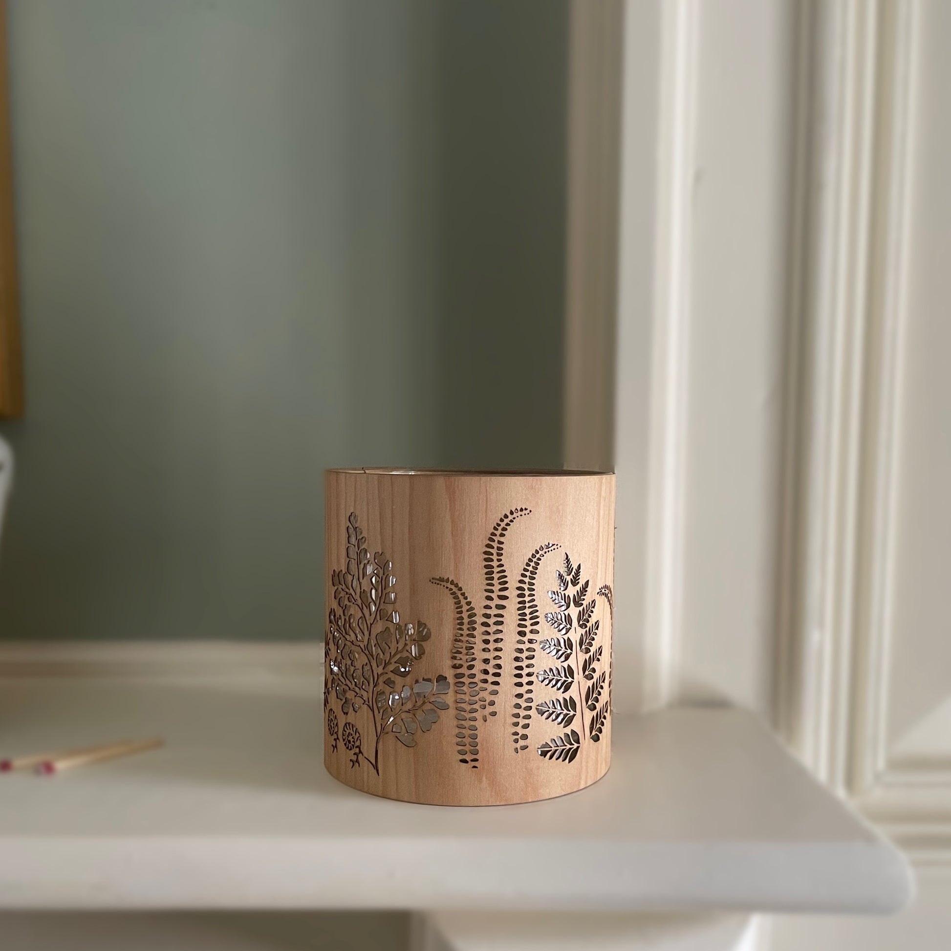 A cylindrical wooden candle holder with laser-cut botanical fern and leaf patterns, sitting on a white mantelpiece or shelf against a muted sage green wall.