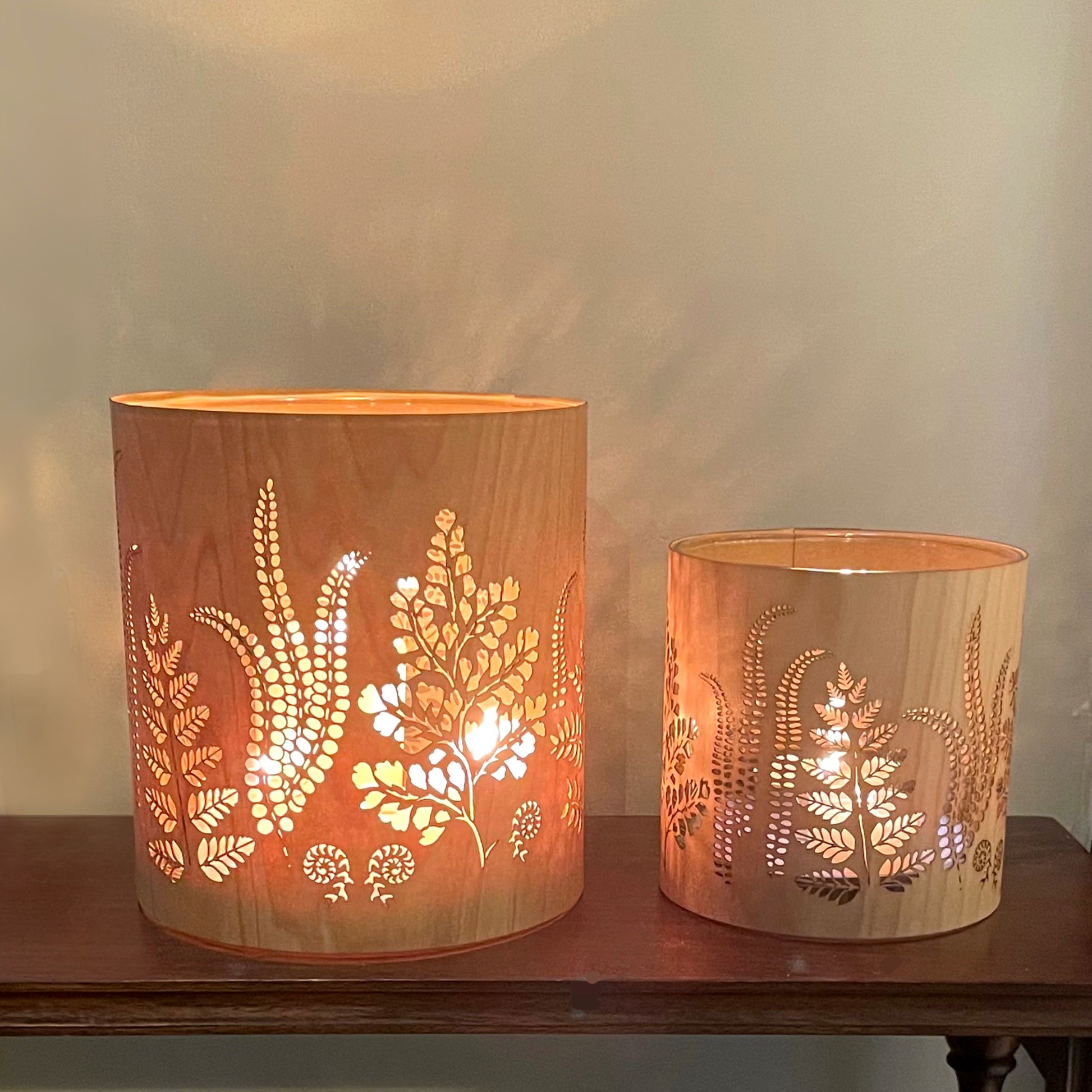 Two wooden decorative candle holders, one large one medium, each with laser-cut fern and leaf patterns, glowing with warm light, displayed on a dark wooden table against a neutral beige wall.