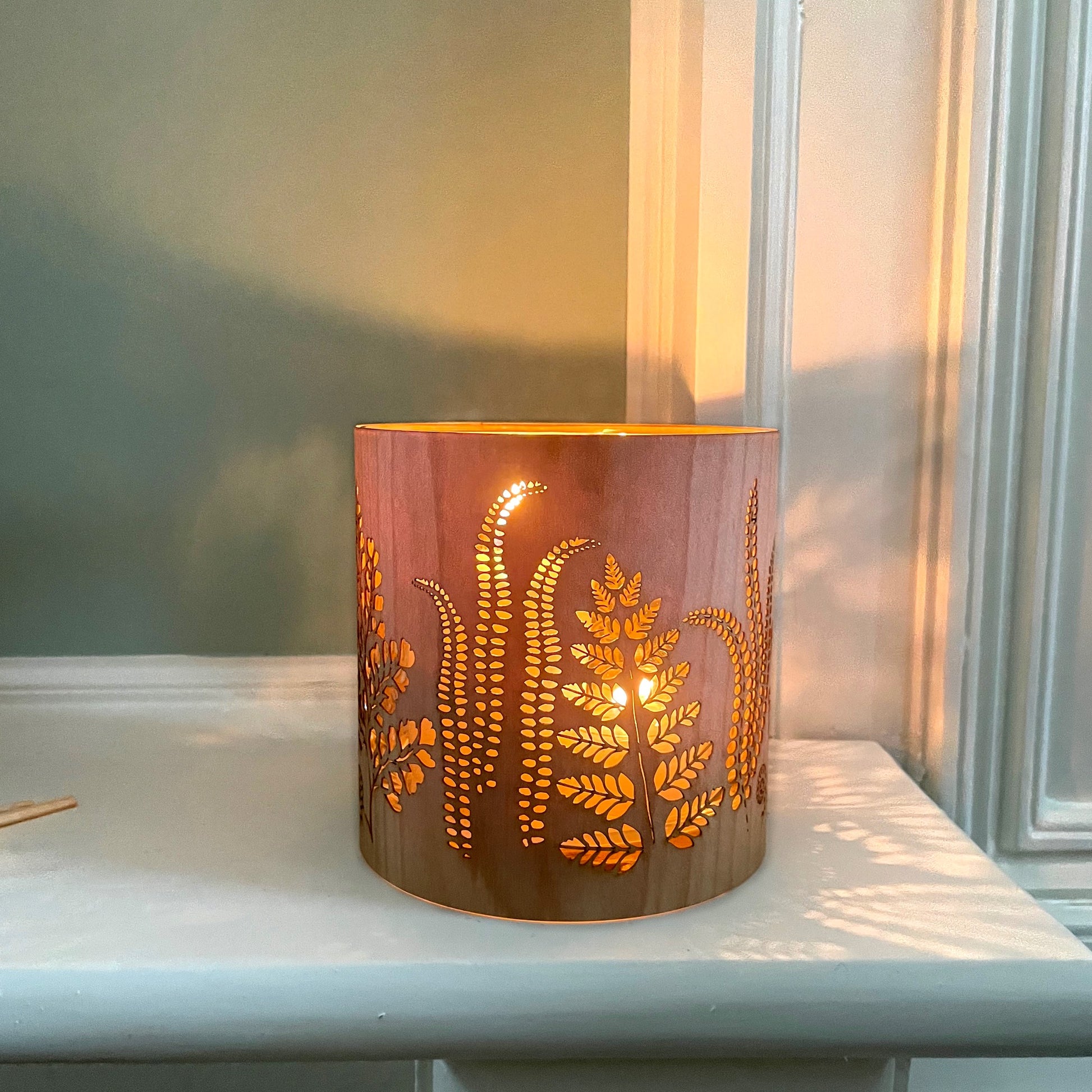 A cylindrical wooden candle holder or lamp with laser-cut fern and leaf patterns, glowing warm orange from an internal light, set on a white windowsill.