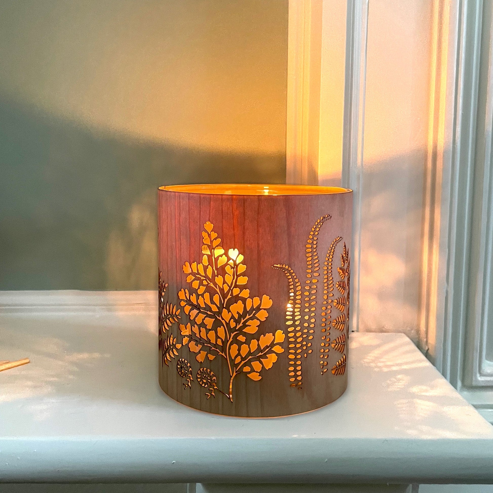 A cylindrical wooden candle holder or lamp with laser-cut botanical leaf patterns, glowing with warm light from a nearby window.