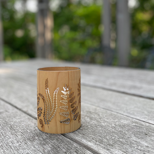 A light natural wood cylindrical cup or candle holder with laser-cut fern patterns, resting on a weathered gray wooden table with a blurred green garden background.