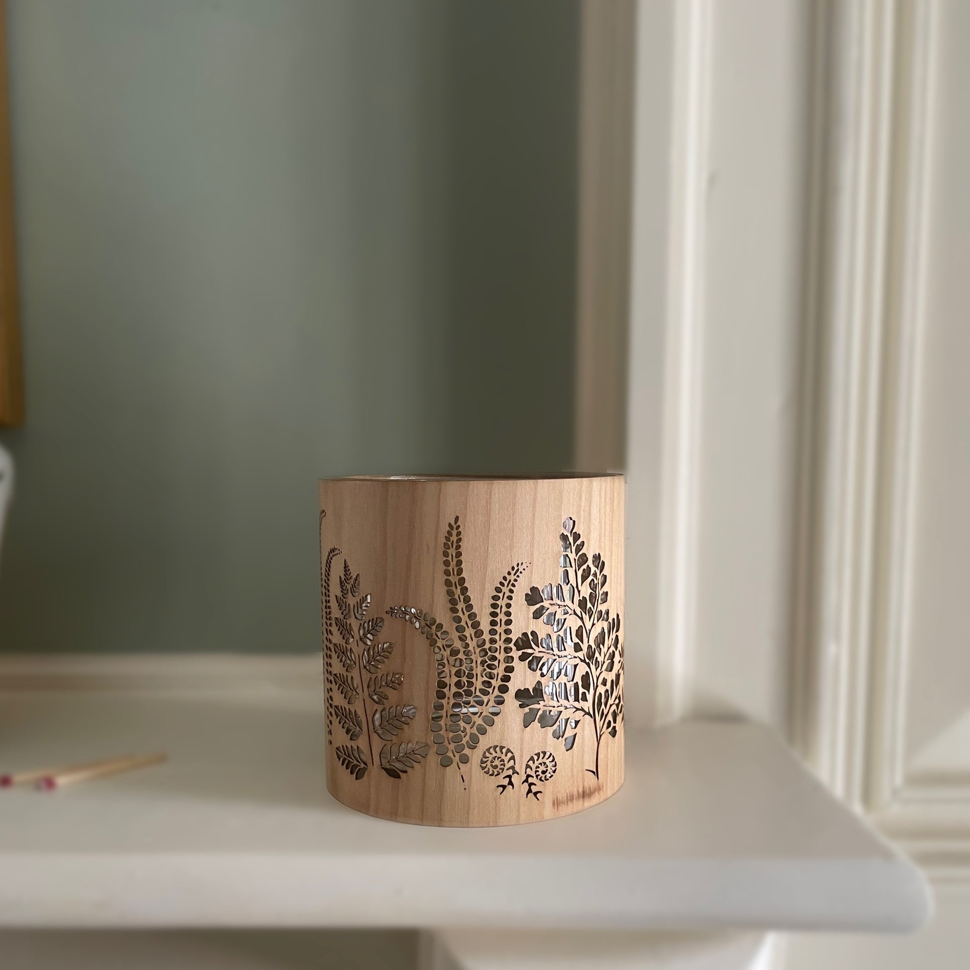 A cylindrical wooden candle holder with a laser-cut botanical fern pattern, sitting on a white mantelpiece or shelf against a muted sage green wall.
