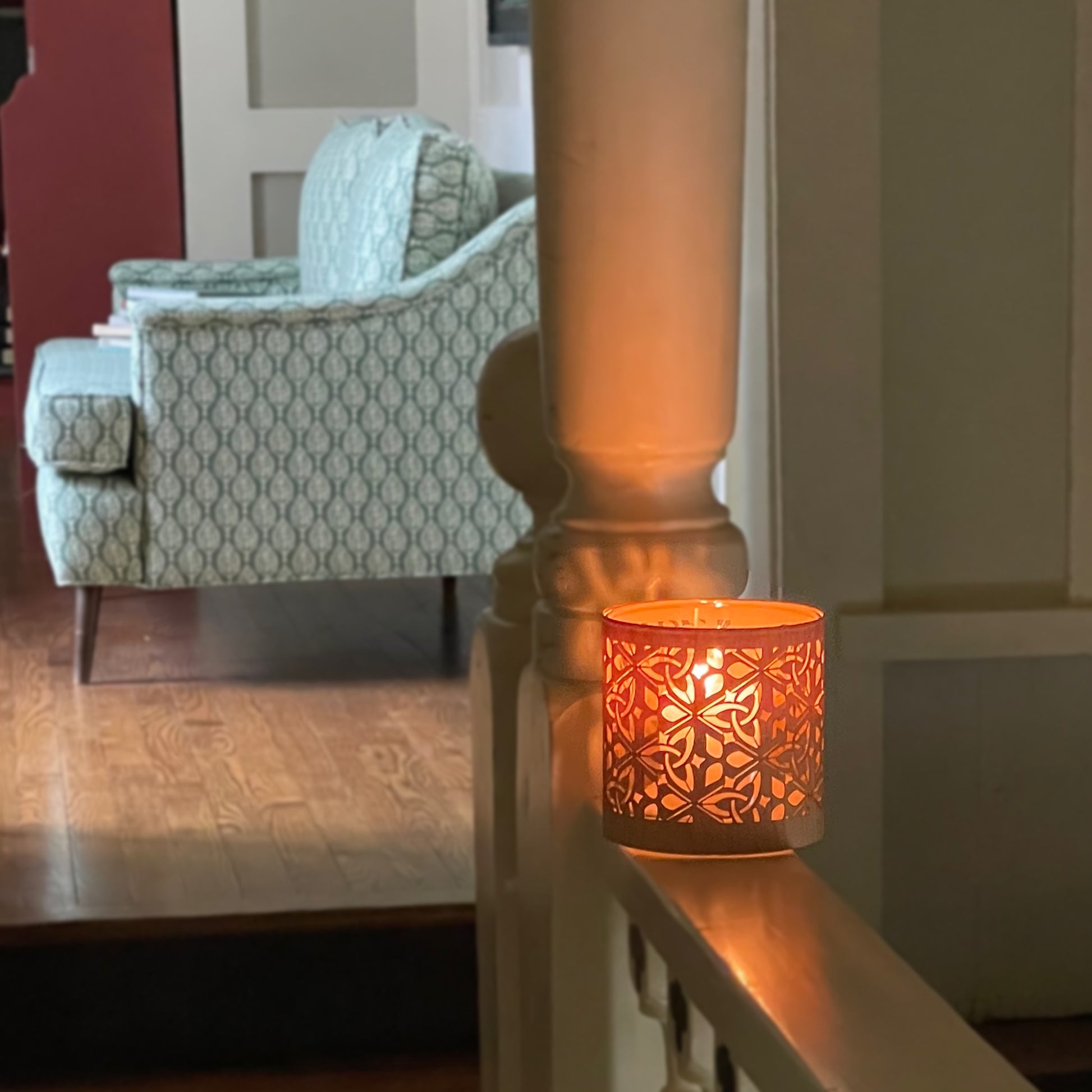 Decorative triquetra candle holder on a railing with a patterned armchair in the background. The latern is made from maple wood veneer and heavy glass. it is lit and casts a warm glow