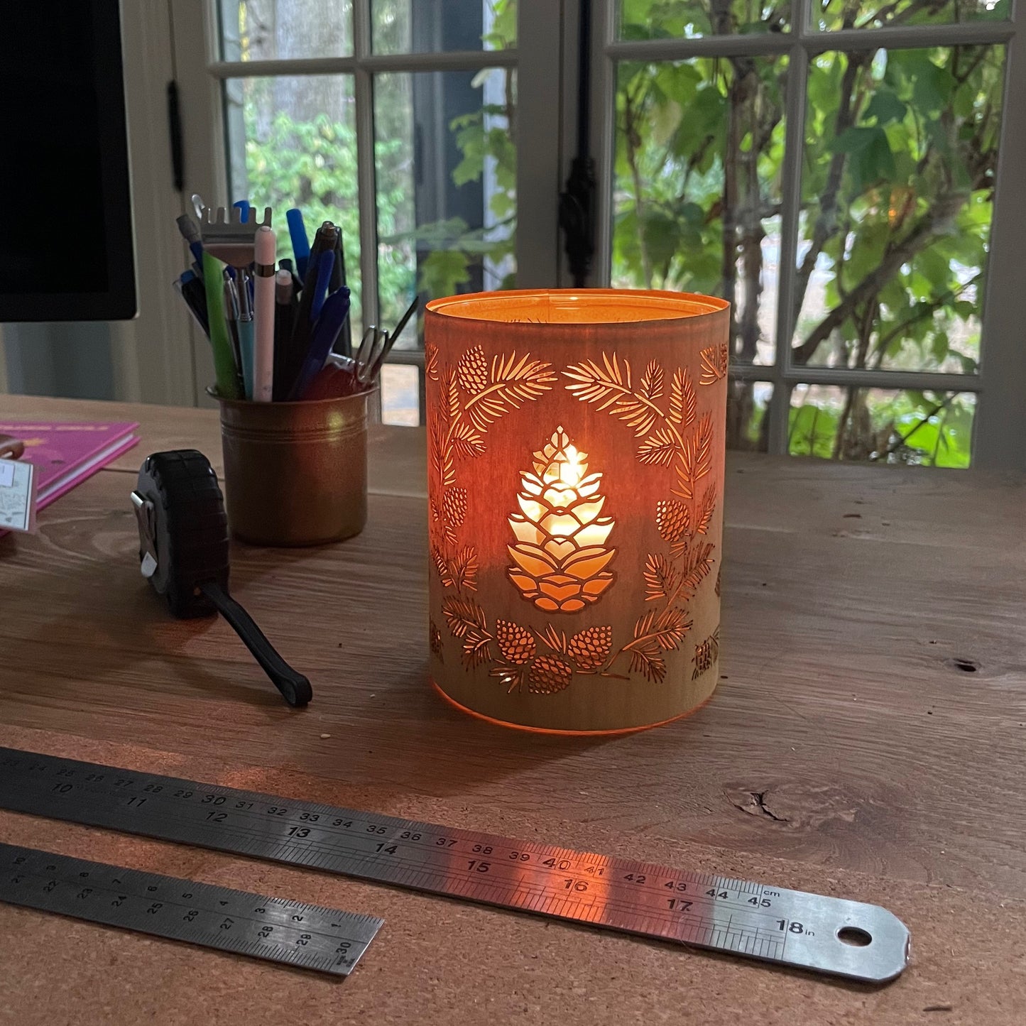 a glass lantern candle holder - size small wrapped in maple veneer with laser cut pinecone design lit with a candle on a desk