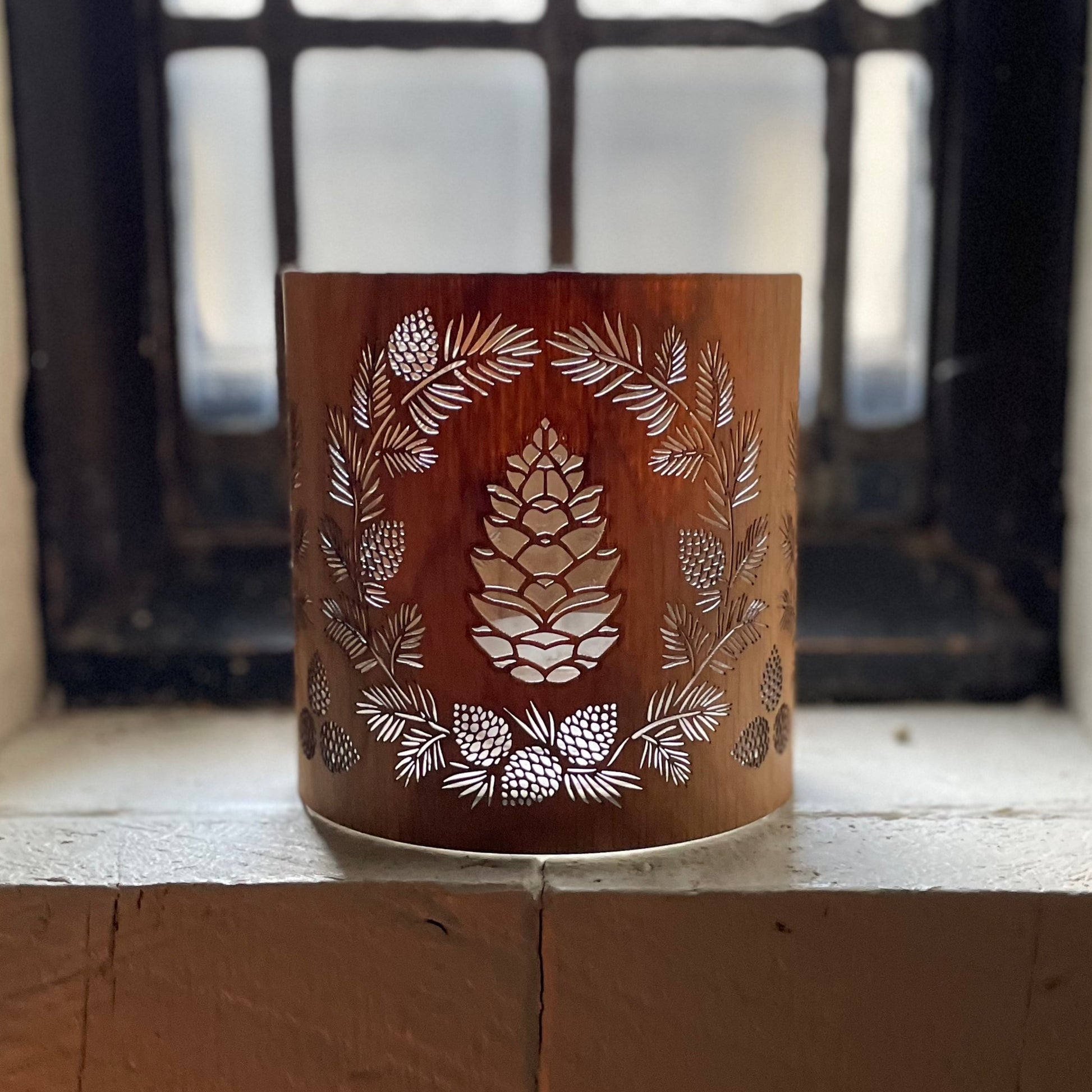 a glass lantern candle holder wrapped in white oak veneer with laser cut pinecone design in a lead glass window with the morning light shining through