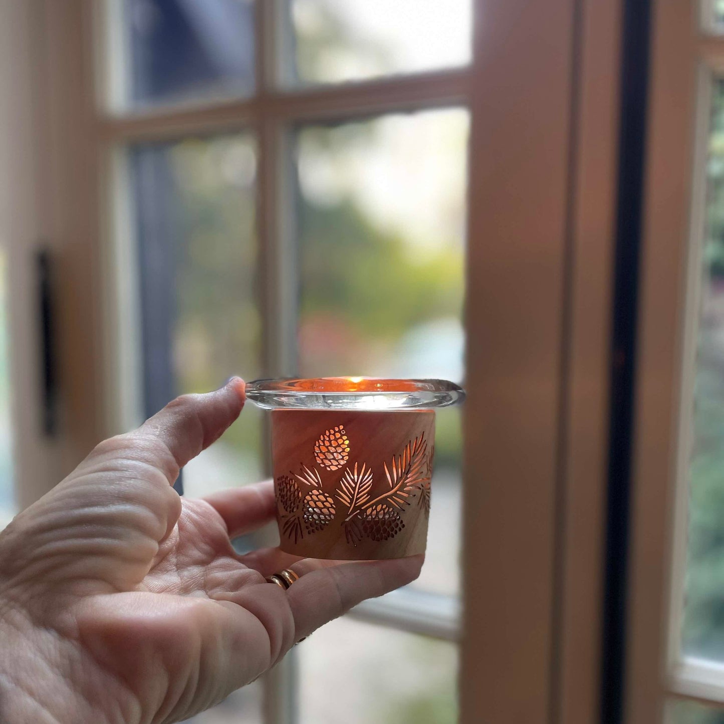 a tealight holder with a pinecone design in wood and glass held in someone’s left hand in front of a window