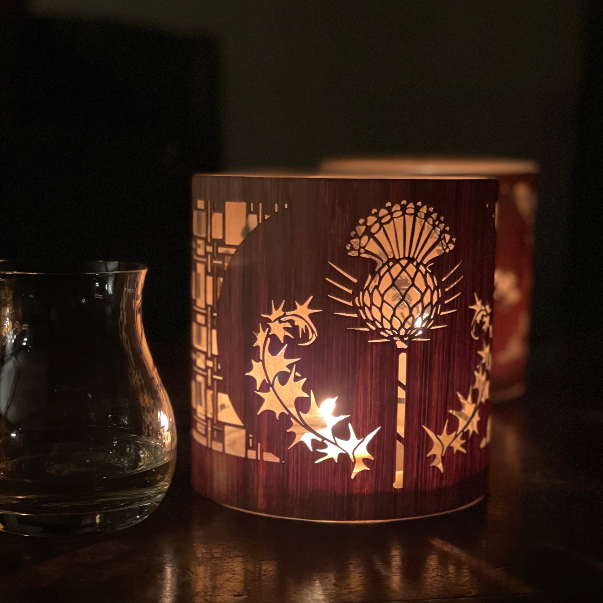 a black walnut lantern with the thistle design lit beside a glass of scotch